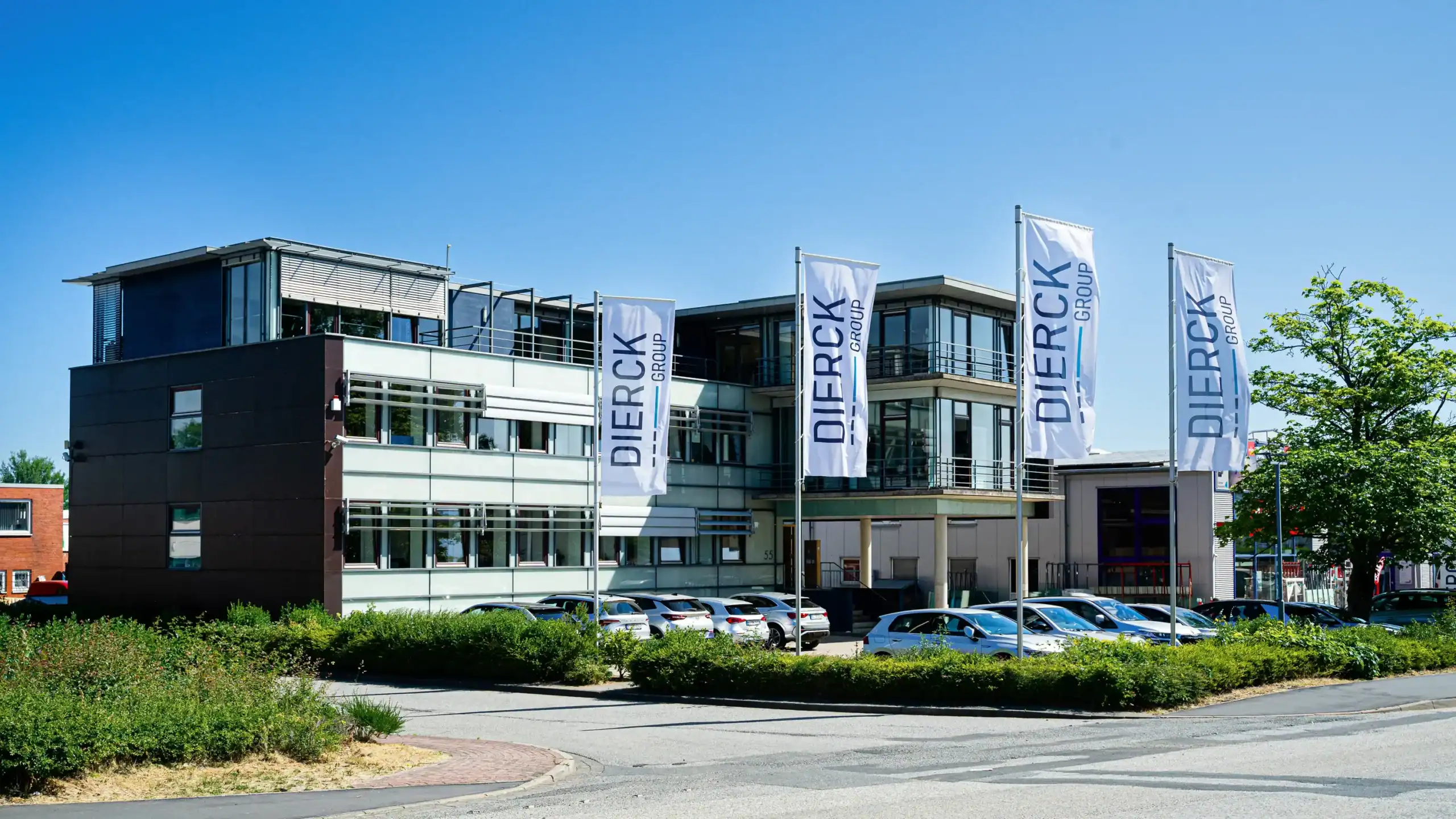 Modernes Bürogebäude der dierckgroup mit vier hohen Fahnen am Eingang, auf denen „dierckgroup“ steht. Vor dem Gebäude mehrere geparkte Autos, umgeben von Grünflächen und Büschen, unter klarem blauem Himmel. Standort Kiel, repräsentativ für IT-Systemhaus und Digitalisierung.