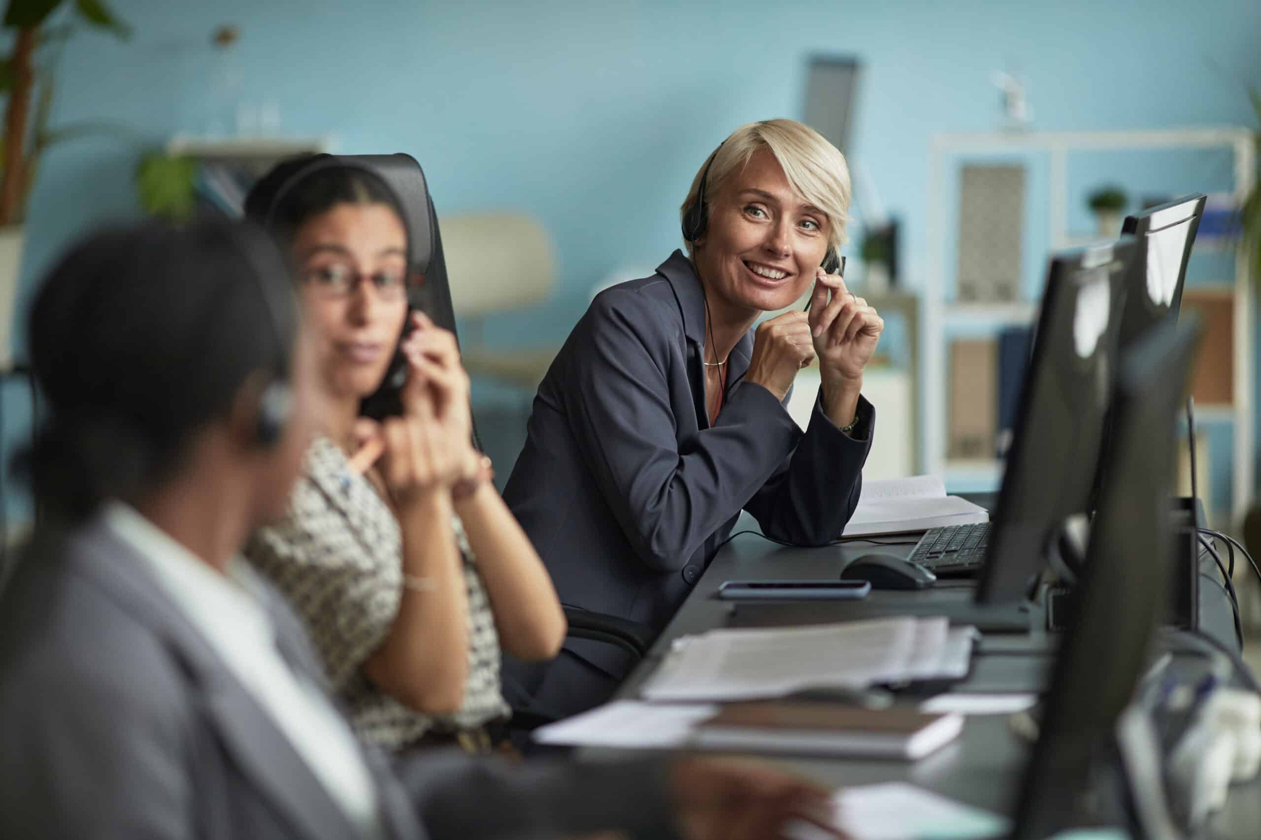 Drei Mitarbeitende in einem modernen Büro mit Headsets sitzen an Schreibtischen vor Computern. Eine lächelnde blonde Frau mittleren Alters im Vordergrund wirkt freundlich und aufmerksam, im Hintergrund zwei jüngere Kolleginnen, die telefonieren. Die Szene vermittelt Kundenservice und IT-Support in einem professionellen Arbeitsumfeld.