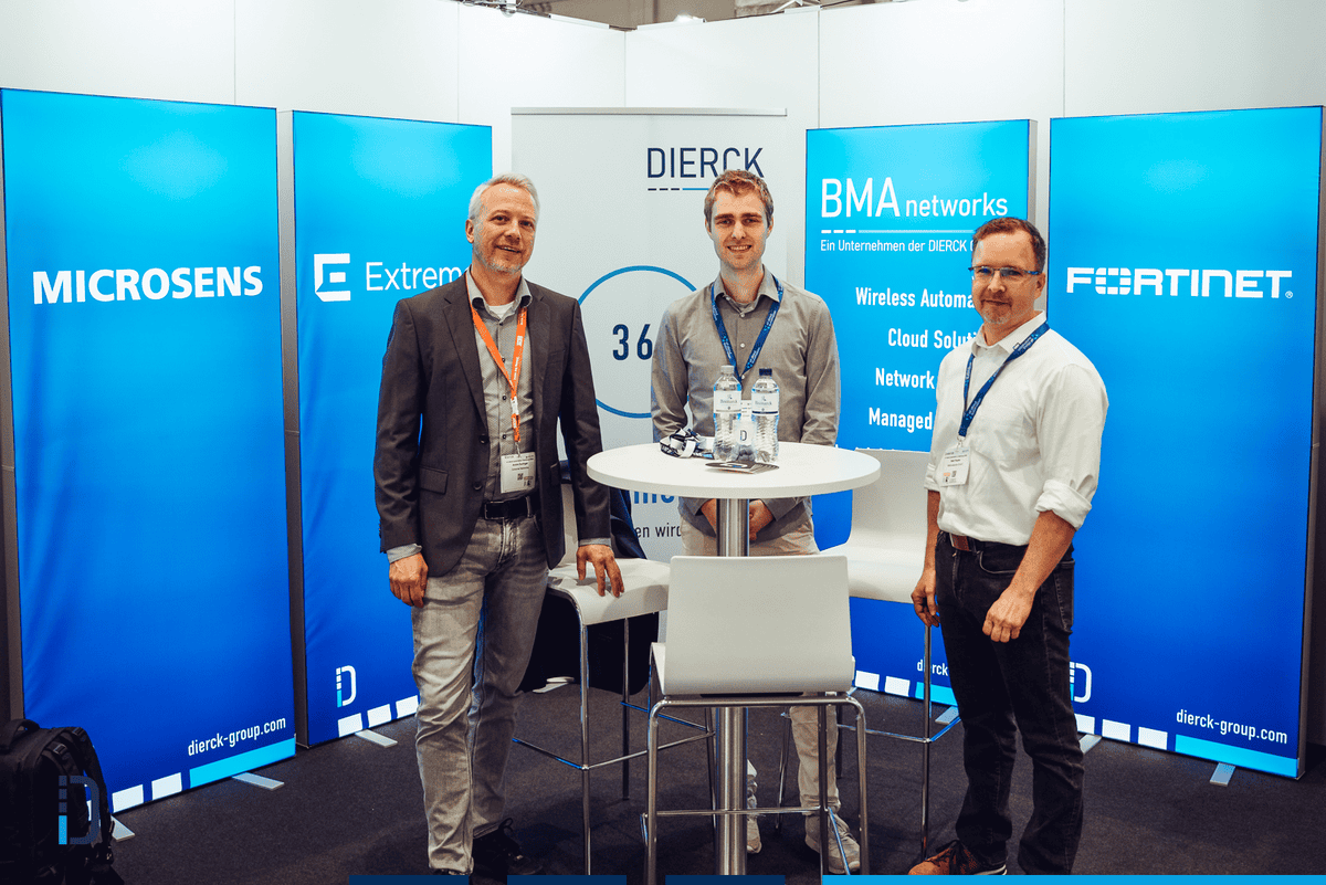 Drei Männer stehen an einem hohen weißen Tisch auf einem Messestand mit blauen Werbebannern im Hintergrund. Die Banner zeigen die Logos und Namen von Microsens, Extreme, BMA networks und Fortinet sowie Hinweise auf Cloud Solutions, Wireless Automation, Network Security und Managed Services. Im Hintergrund ist ein Banner mit dem Schriftzug dierckgroup und dem Slogan „360° IT Solutions“ zu sehen. Die Männer tragen Business-Kleidung und Namensschilder.