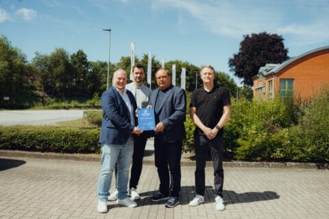 Vier Männer stehen vor einem Firmengebäude im Freien auf gepflastertem Boden. Zwei von ihnen halten gemeinsam eine blaue Urkunde mit dem Schriftzug „dierckgroup“ und weiteren Texten. Im Hintergrund sind Fahnenmasten mit dem Logo der dierckgroup sowie grüne Büsche und Bäume zu sehen. Die Szene vermittelt einen professionellen, freundlichen Moment im Bereich IT-Systemhaus und Managed Services.
