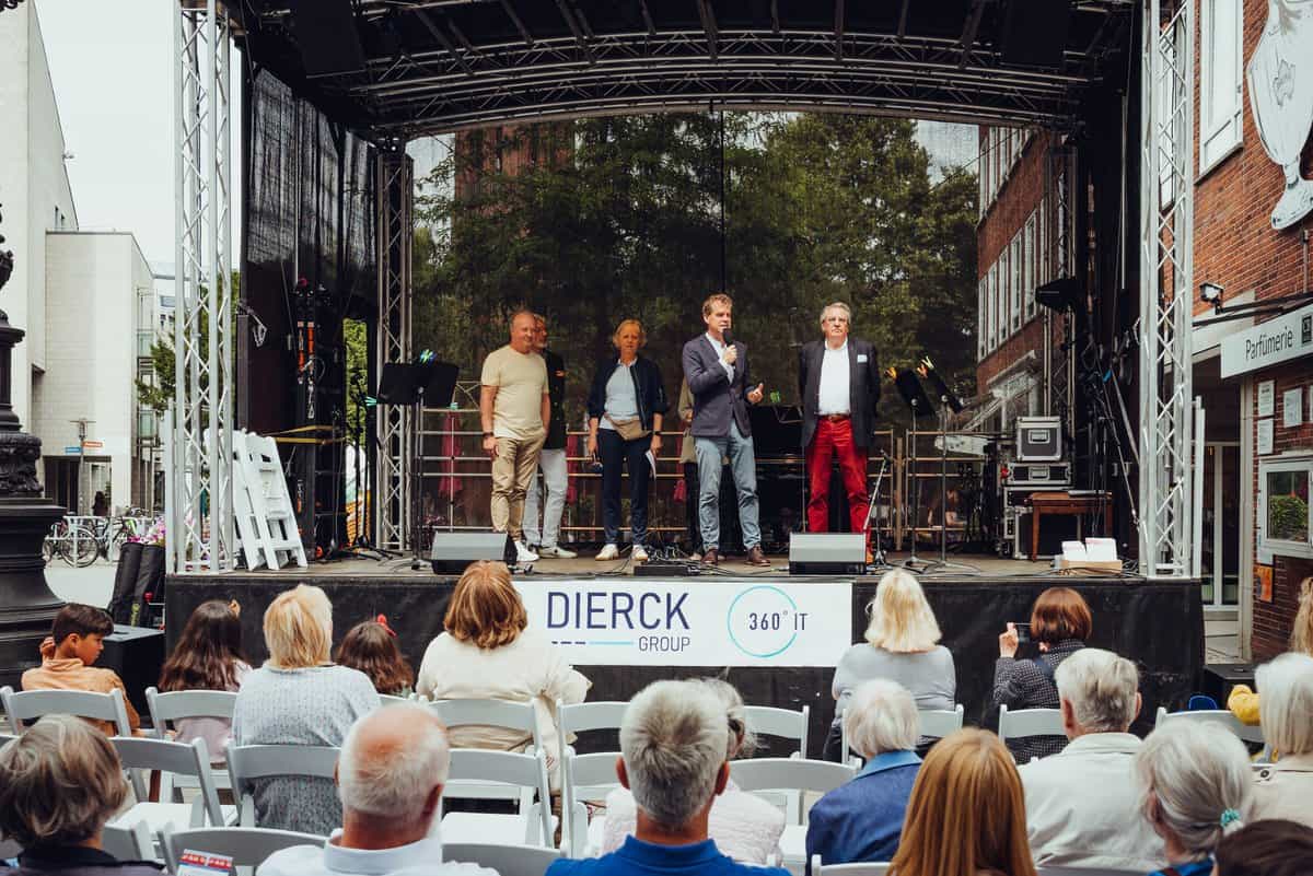 Vier Personen stehen auf einer überdachten Bühne im Freien, eine davon spricht in ein Mikrofon. Vor der Bühne sitzen mehrere Zuschauer auf weißen Stühlen. An der Bühnenfront ist ein Banner mit dem Schriftzug „dierckgroup 360° IT“ angebracht. Im Hintergrund sind Bäume und Gebäude zu sehen. Die Szene vermittelt eine Veranstaltung oder Präsentation, passend zu IT-Lösungen und Managed Services der dierckgroup.