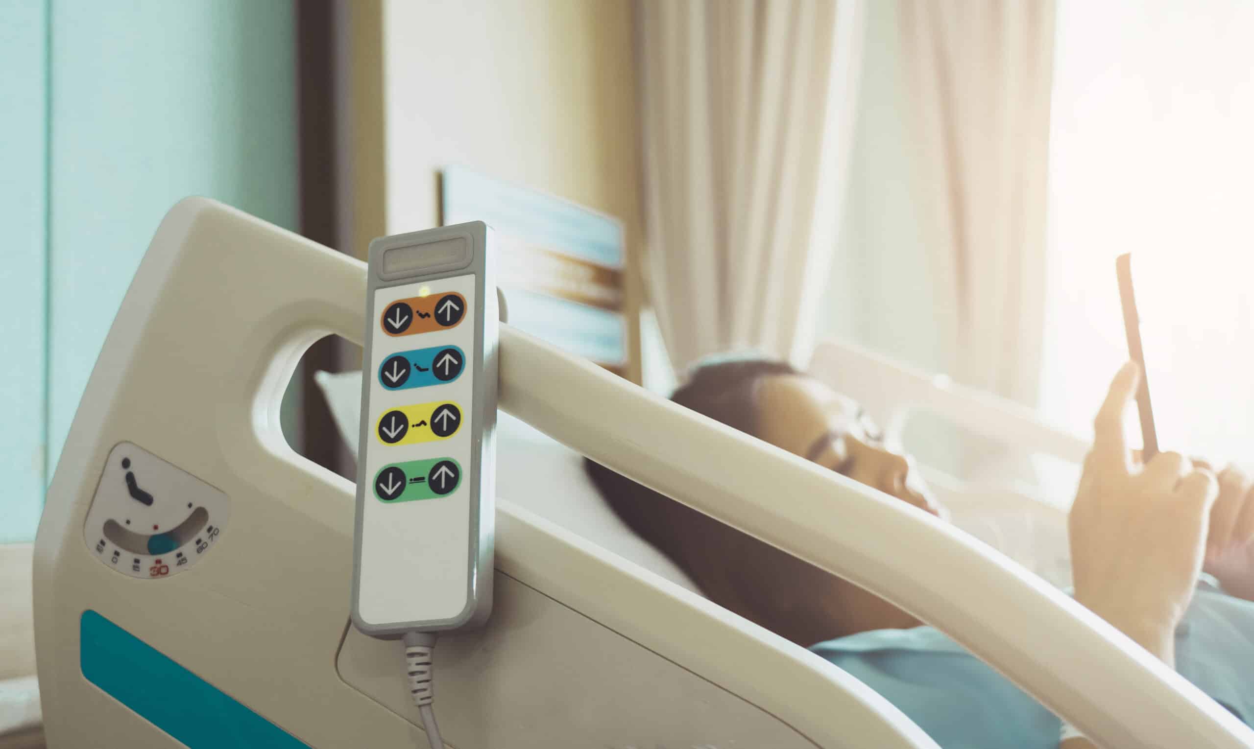 Fernbedienung für Krankenbett mit Pfeiltasten zur Verstellung, hängt am Bettgeländer eines hellen Patientenzimmers, im Hintergrund liegt eine Frau im Bett und hält ein Smartphone, sanfte Beleuchtung durch Tageslicht.