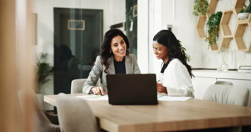 Zwei Frauen sitzen an einem Holztisch in einem modernen, hellen Büro und schauen gemeinsam auf einen Laptop, dabei lächeln sie und wirken engagiert in einem Beratungsgespräch. Im Hintergrund sind Pflanzen und dekorative Wandregale mit sechseckigen Fächern zu sehen.