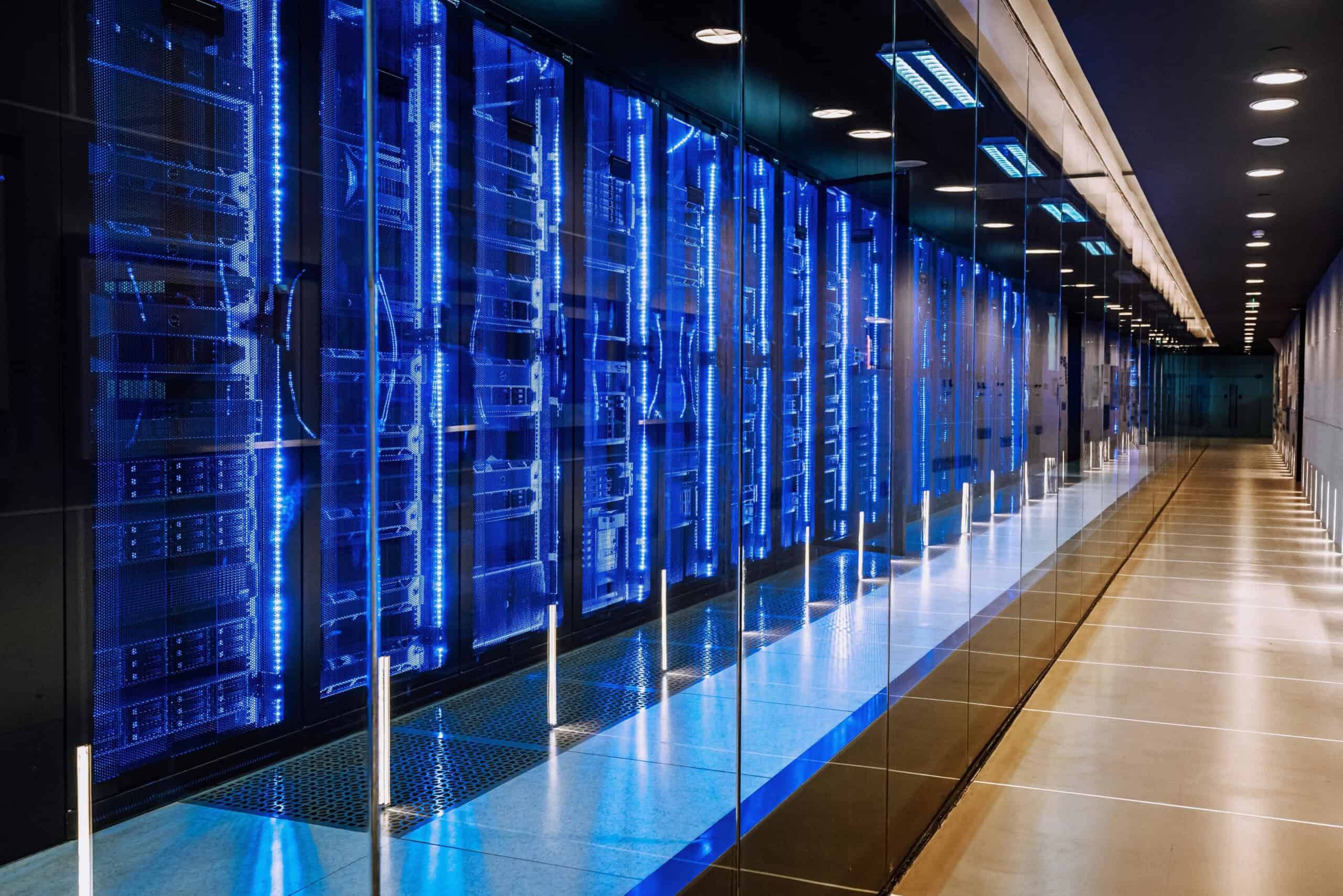 Langer, moderner Serverraum mit mehreren Racks, die mit blau leuchtenden Netzwerk- und Datacenter-Komponenten bestückt sind, reflektiert in einer Glaswand, symbolisiert IT Solutions, Managed Services und Cloud Services der dierckgroup.