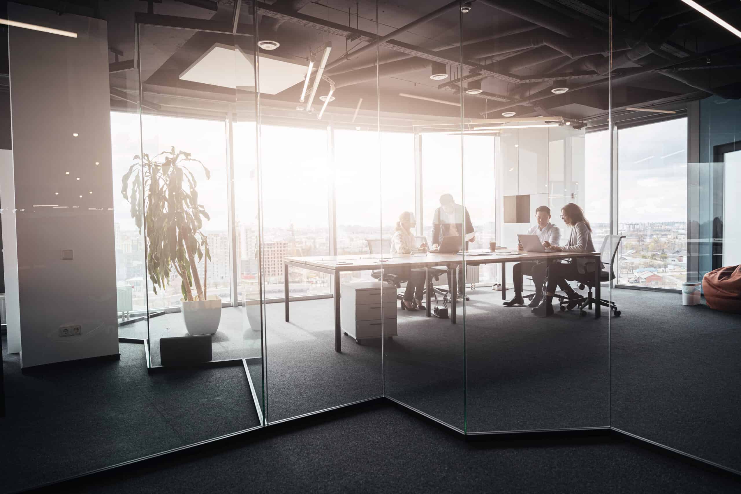 Vier Geschäftsleute sitzen und stehen an einem großen Tisch in einem modernen Büro mit bodentiefen Fenstern und Blick auf die Stadt. Im Raum sind ein großer Pflanzenkübel, ein Sitzsack und Bürostühle zu sehen. Die Atmosphäre wirkt professionell und kooperativ.