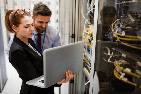 Zwei Techniker, eine Frau und ein Mann, analysieren gemeinsam Daten auf einem Laptop vor einem Serverrack mit zahlreichen vernetzten Kabeln in einem Rechenzentrum, symbolisiert IT-Support und Netzwerklösungen der dierckgroup.