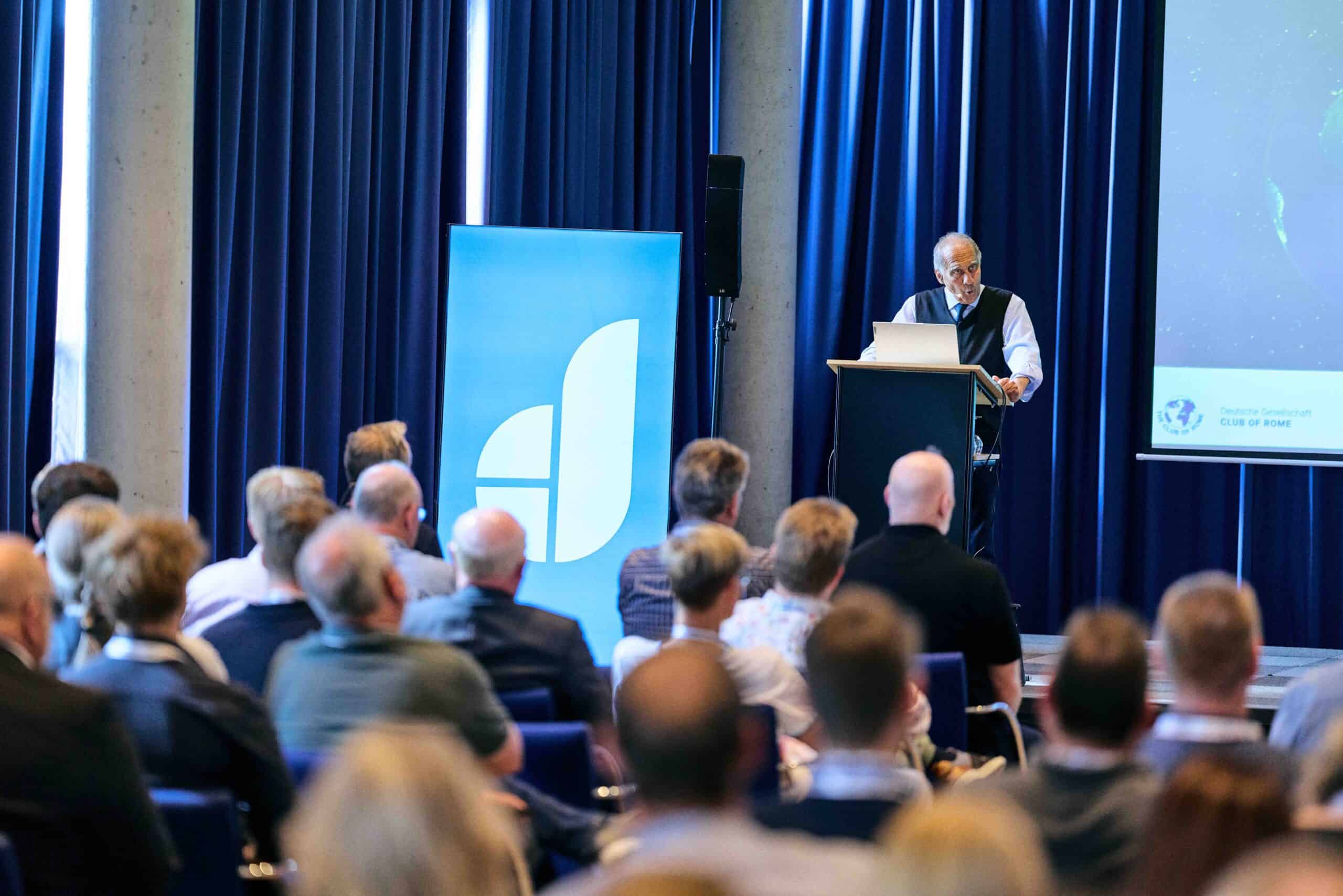 Mann hält Vortrag vor Publikum in einem Konferenzraum mit blauem Vorhang, neben ihm steht ein Banner mit stilisiertem weißem Logo auf blauem Hintergrund, rechts ist eine Projektionsfläche mit teilweise sichtbarem Globus und Text, Zuschauer sitzen in Reihen und hören aufmerksam zu.