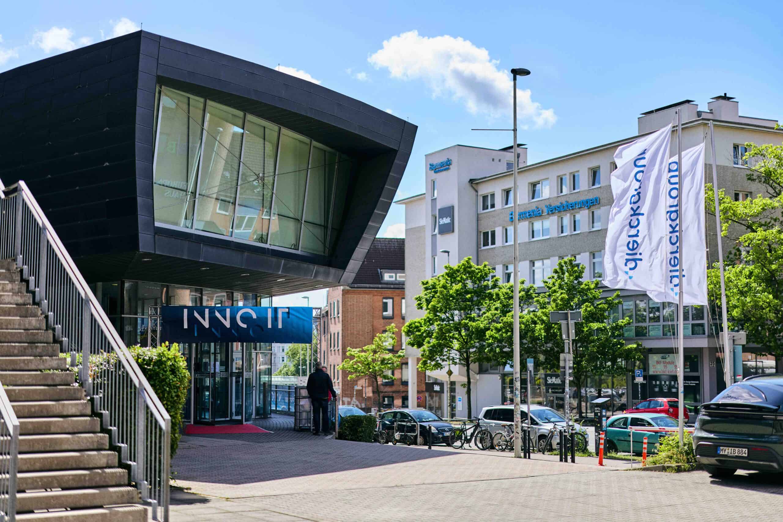 Modernes Bürogebäude mit großem, schräg auskragendem Glasfenster und blauem Eingangsschild „INNO IT“, davor Gehweg mit Treppe links, zwei wehenden Fahnen mit „dierckgroup“ Schriftzug rechts, mehrere geparkte Autos und grüne Bäume entlang der Straße bei sonnigem Wetter.