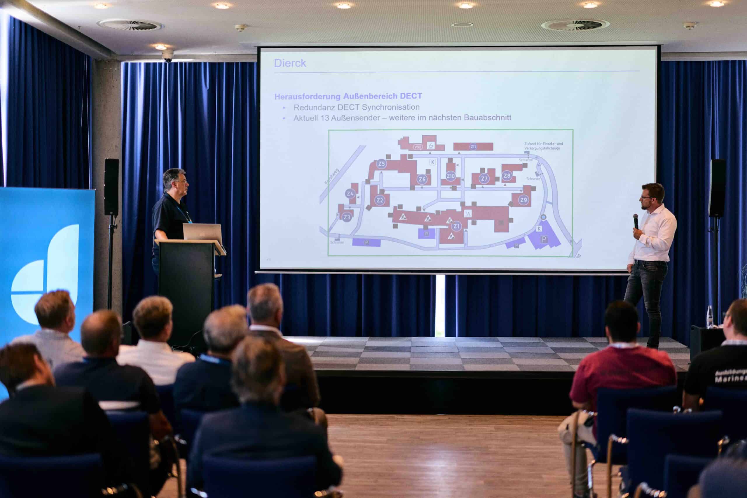 Zwei Männer präsentieren vor Publikum in einem Konferenzraum eine Folie mit dem Titel „Herausforderung Außenbereich DECT“ und einer schematischen Karte eines Gebäudekomplexes. Im Hintergrund dunkelblaue Vorhänge, links ein blaues Banner mit weißem Logo. Die Szene vermittelt eine professionelle IT-Beratung oder Fachvortrag.