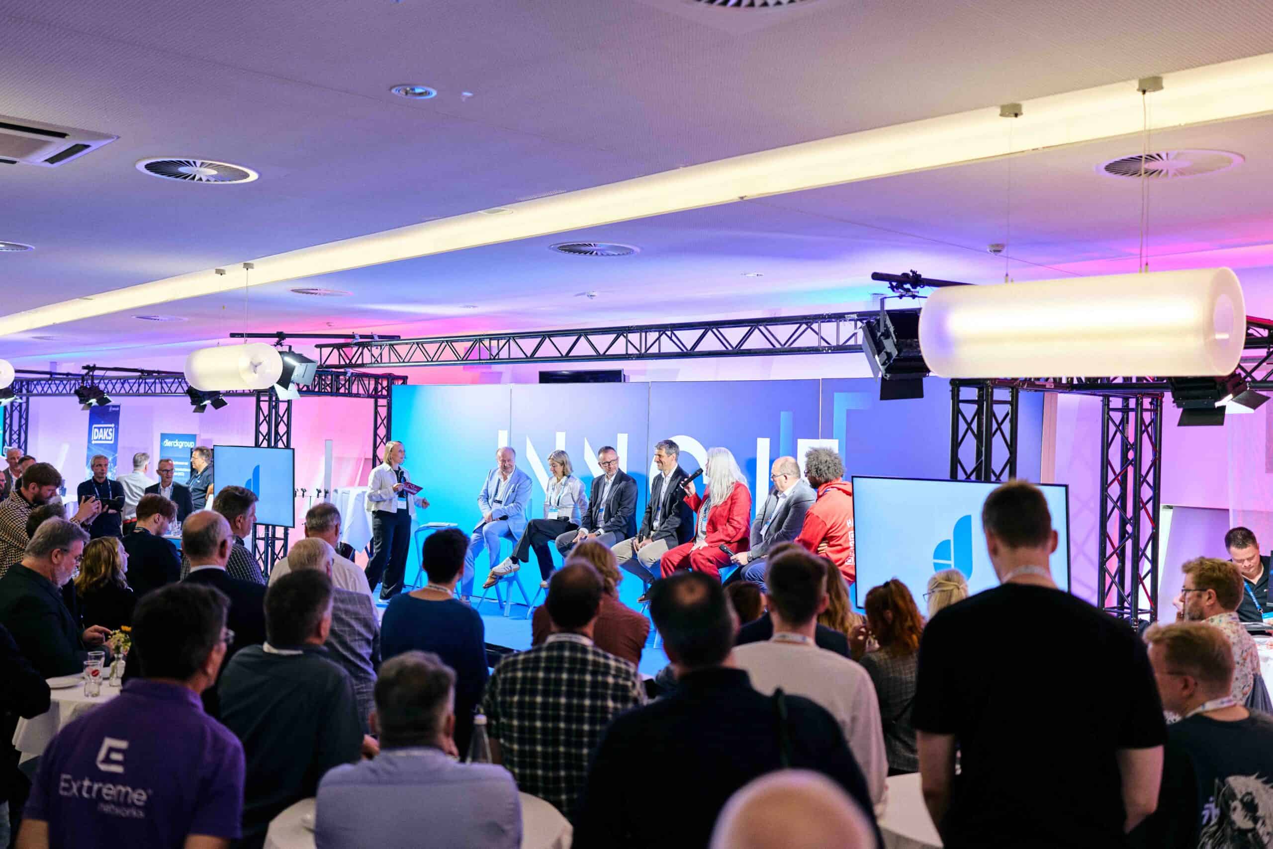 Podiumsdiskussion mit acht Personen auf einer Bühne vor Publikum in einem modernen Veranstaltungsraum, beleuchtet mit blauem und rosa Licht, zwei große Bildschirme zeigen Grafiken, Teilnehmer tragen Business- und Casual-Kleidung, Veranstaltung wirkt professionell und thematisch passend zu IT-Lösungen und Digitalisierung.