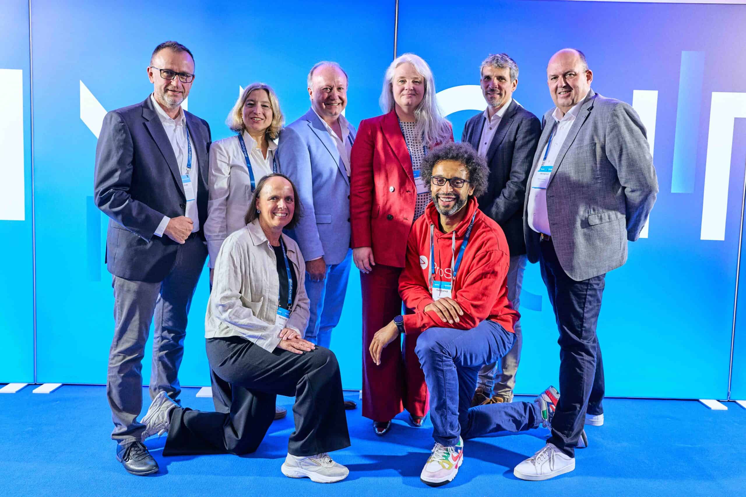 Acht Personen in Business- und Freizeitkleidung posieren vor einer hellblauen Wand mit teilweise sichtbarem, großem Schriftzug. Zwei Personen knien vorne, sechs stehen dahinter. Die Gruppe wirkt freundlich und professionell, passend zu einem IT-Systemhaus-Umfeld.