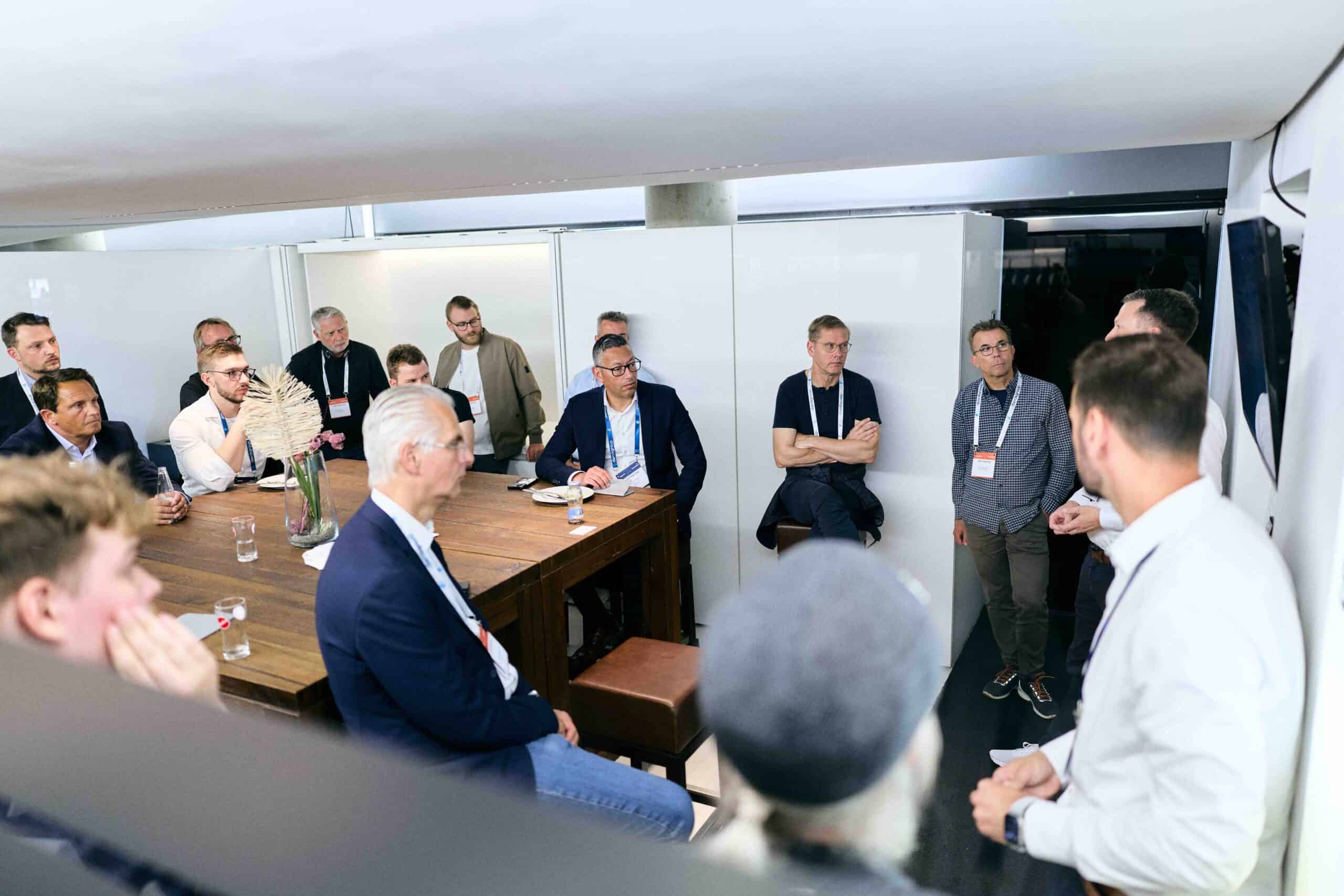 Mehrere Männer unterschiedlichen Alters sitzen und stehen in einem modernen, hellen Besprechungsraum um einen großen Holztisch, hören aufmerksam einem stehenden Sprecher zu, der eine Präsentation hält. Die Atmosphäre wirkt professionell und konzentriert.