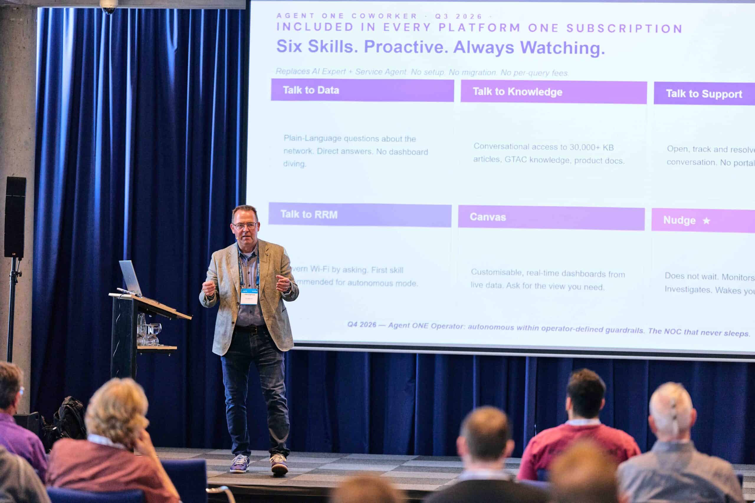 Mann in grauem Sakko und Jeans hält Präsentation vor Publikum in Konferenzraum mit blauem Vorhang, auf großer Leinwand sind Stichpunkte zu Plattform-Funktionen wie „Talk to Data“, „Talk to Knowledge“ und „Talk to Support“ zu sehen, Szene vermittelt IT-Beratung und Digitalisierung.
