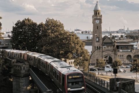 Vorbeifahrende S-Bahn auf erhöhter Strecke neben dem historischen St.-Pauli-Landungsbrücken-Turm mit Uhr, im Hintergrund Bäume, Hafenanlagen und Wasser, bei bewölktem Himmel.