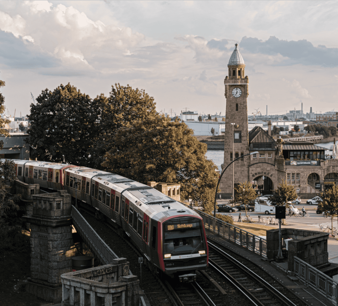 Vorbeifahrende S-Bahn auf erhöhter Strecke neben dem historischen St.-Pauli-Landungsbrücken-Turm mit Uhr, im Hintergrund Bäume, Hafenanlagen und Wasser, bei bewölktem Himmel.
