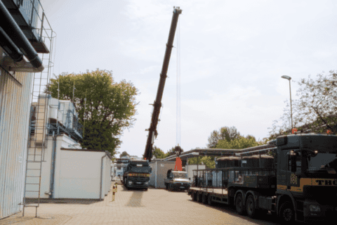 Hof eines Industriegebäudes mit zwei großen Lastwagen und einem Kran, der eine schwere Last an einem Haken hebt, umgeben von Bäumen und Gebäudeteilen, bei bewölktem Himmel.
