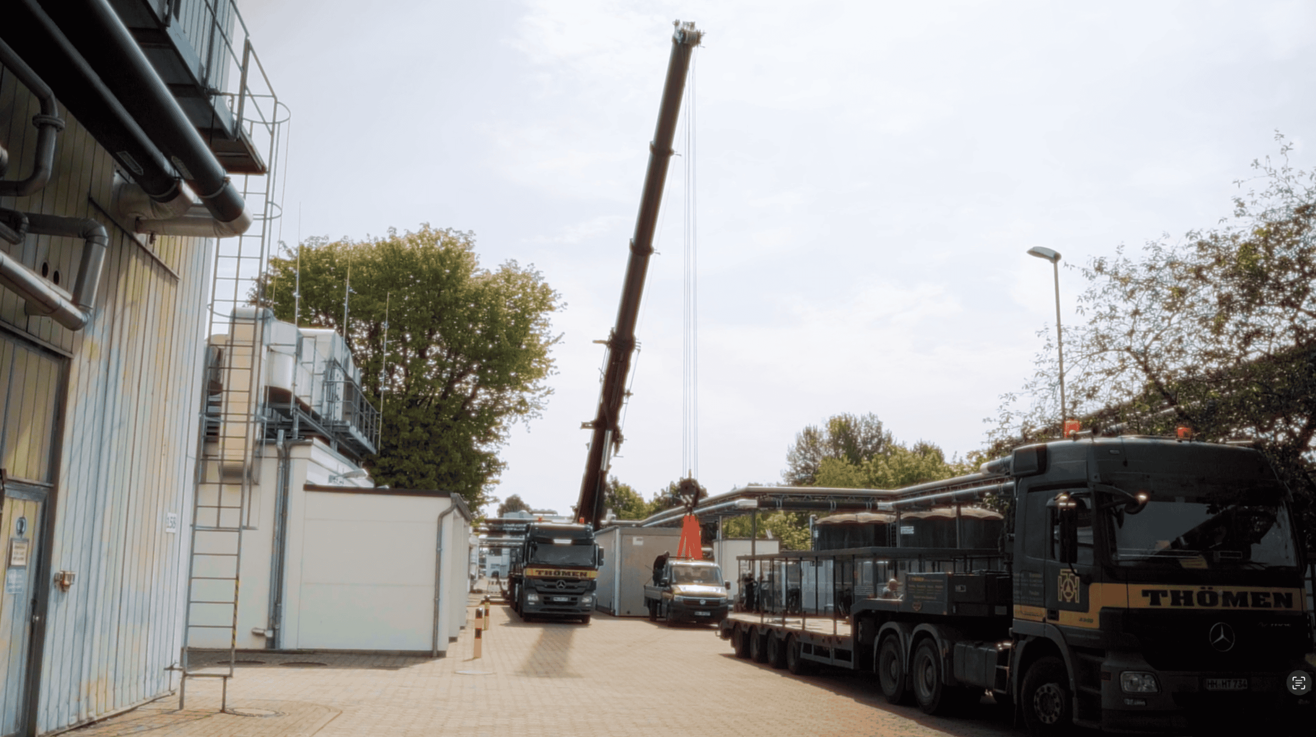 Hof eines Industriegebäudes mit zwei großen Lastwagen und einem Kran, der eine schwere Last an einem Haken hebt, umgeben von Bäumen und Gebäudeteilen, bei bewölktem Himmel.