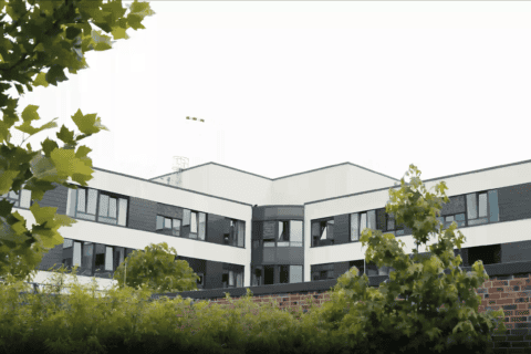 Modernes Bürogebäude mit weißen und dunklen Fassadenelementen, umgeben von grünen Bäumen und Sträuchern, aufgenommen bei bewölktem Himmel.