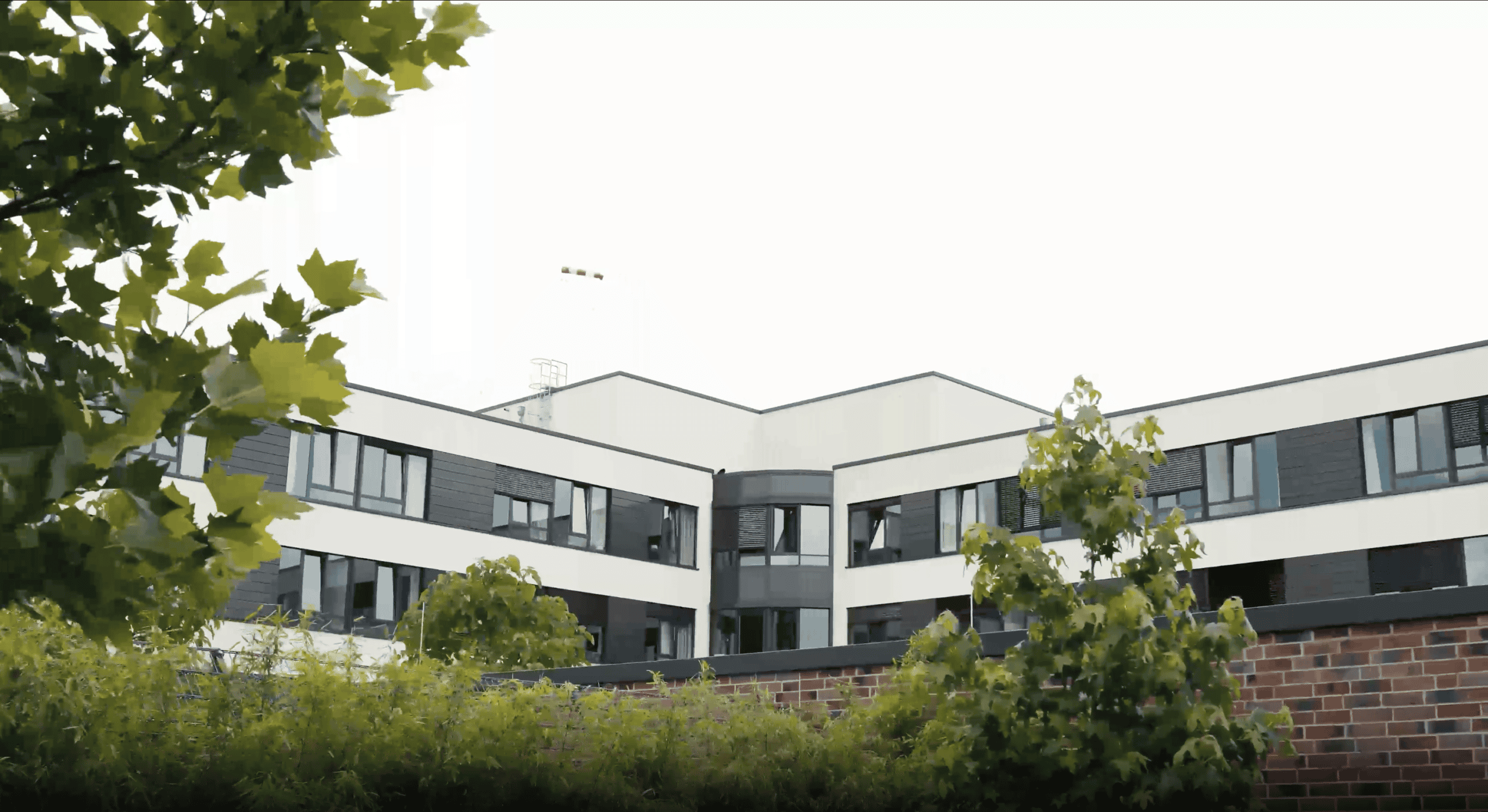 Modernes Bürogebäude mit weißen und dunklen Fassadenelementen, umgeben von grünen Bäumen und Sträuchern, aufgenommen bei bewölktem Himmel.