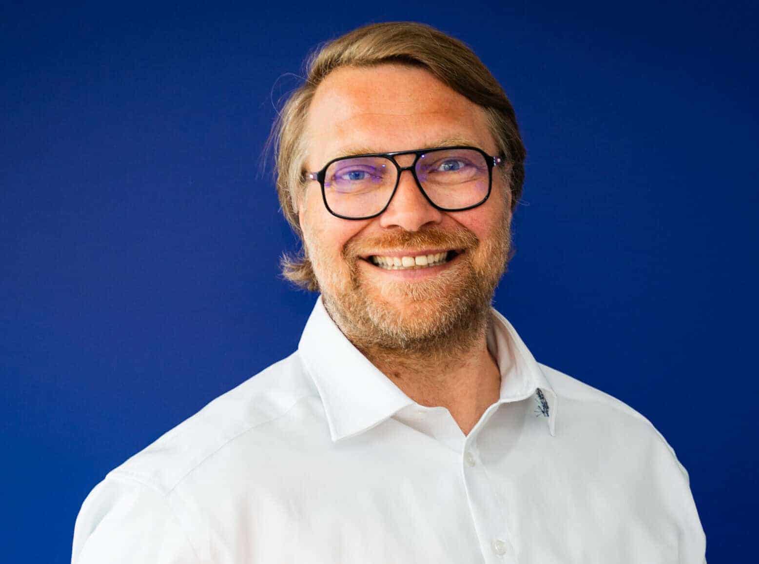 Mann mit Brille und weißem Hemd lächelt vor blauem Hintergrund und hält ein großes oranges Stofftier in Form eines Fisches.