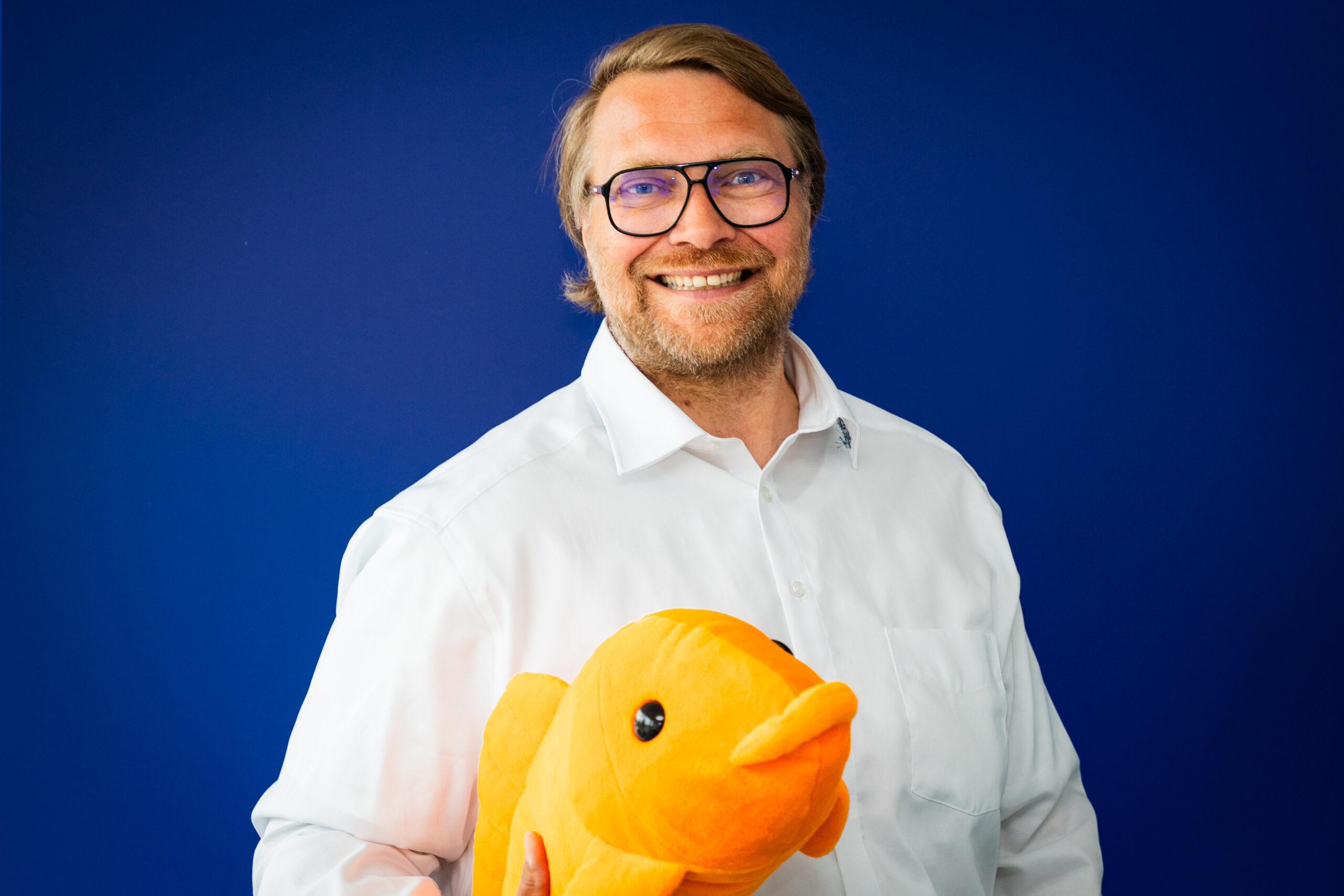 Mann mit Brille und weißem Hemd lächelt vor blauem Hintergrund und hält ein großes oranges Stofftier in Form eines Fisches.