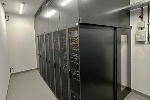 Serverraum mit mehreren schwarzen Serverracks, in denen Kabel und IT-Hardware sichtbar sind, helle Beleuchtung, weiße Wände, sauberer und ordentlicher Raum, typisches Datacenter-Setup für IT- und Cloud-Services.