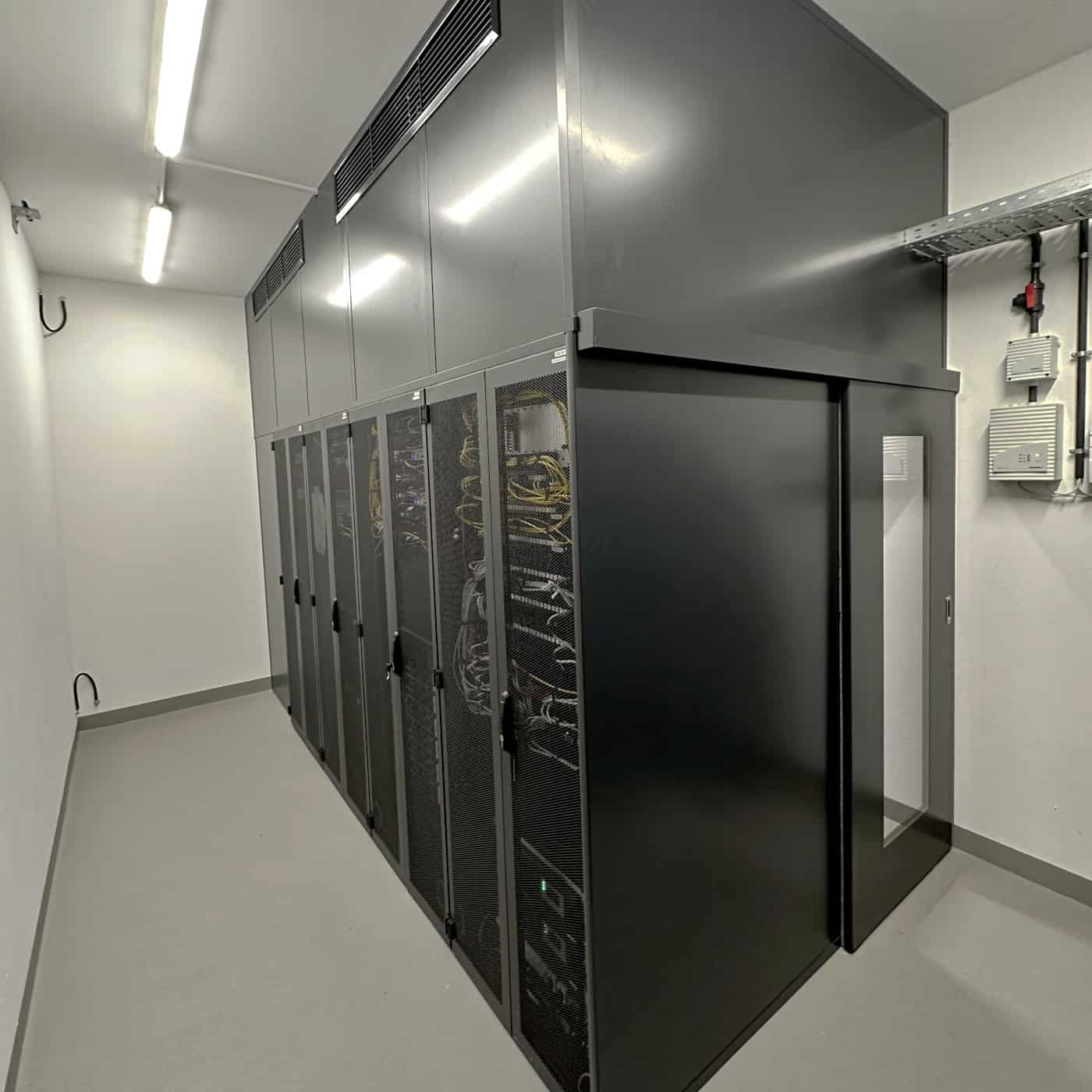 Serverraum mit mehreren schwarzen Serverracks, in denen Kabel und IT-Hardware sichtbar sind, helle Beleuchtung, weiße Wände, sauberer und ordentlicher Raum, typisches Datacenter-Setup für IT- und Cloud-Services.