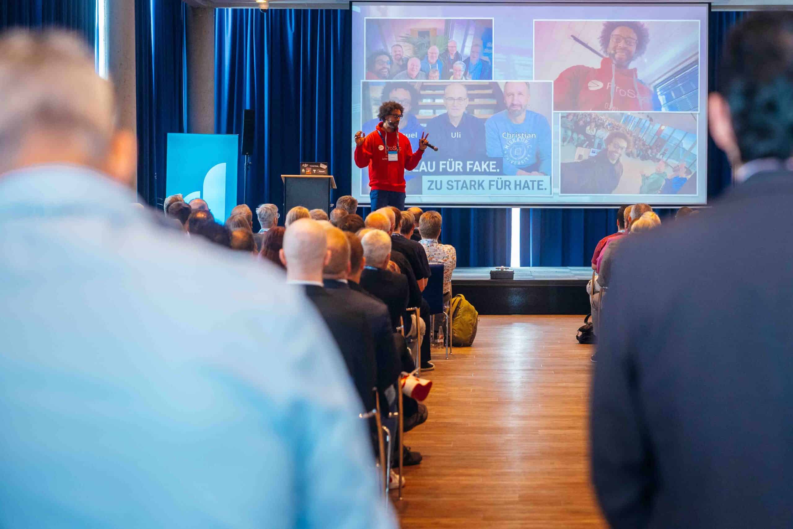 Vortragender mit rotem Pullover spricht vor Publikum in einem Konferenzraum, im Hintergrund große Leinwand mit mehreren Porträtfotos und Textfragment „...AU FÜR FAKE. ... ZU STARK FÜR HATE.“ Publikum sitzt auf Stühlen, Raum mit Holzboden und blauen Vorhängen, Veranstaltung im Kontext von IT-Lösungen und Digitalisierung.