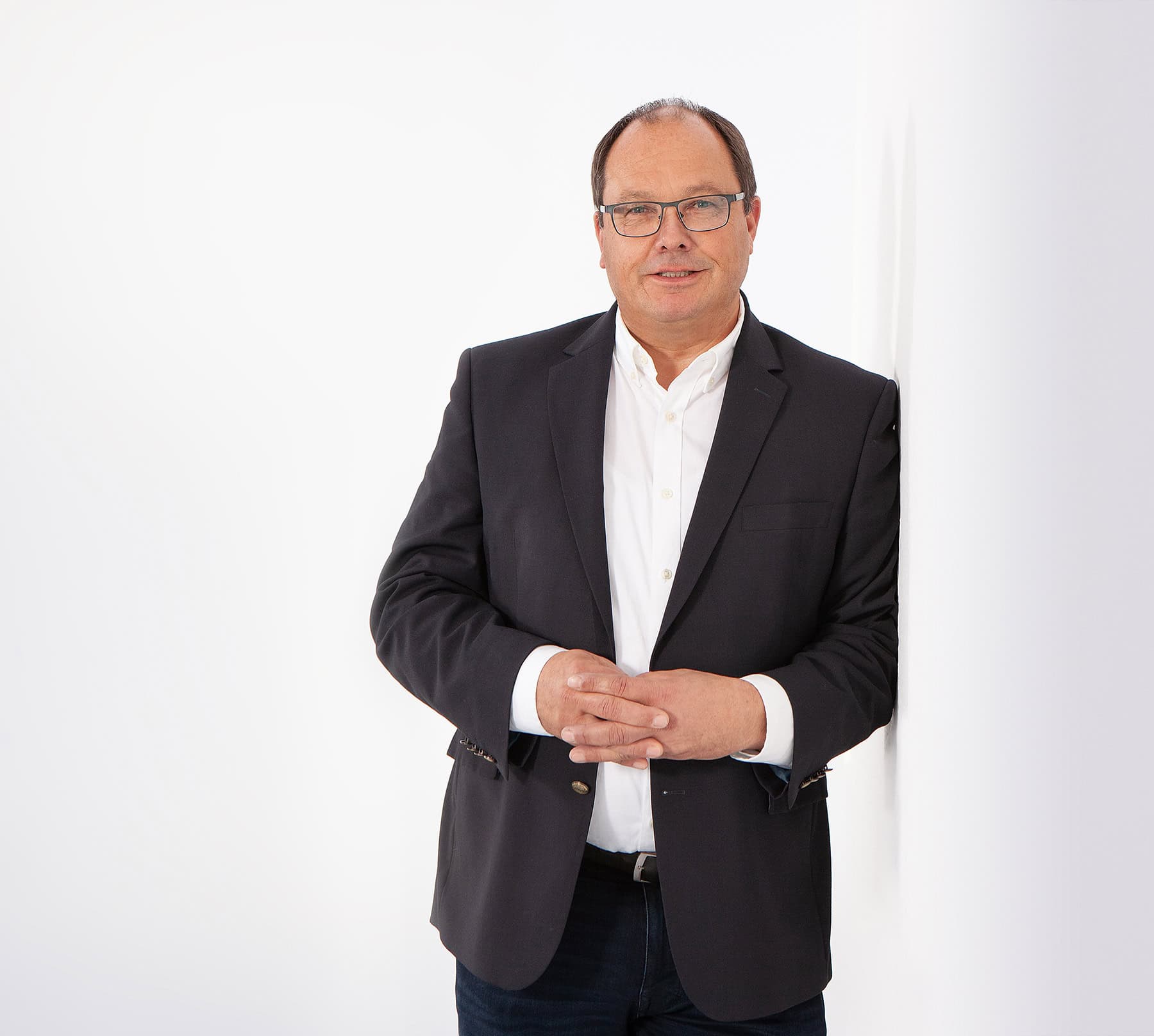 Mann mittleren Alters mit Brille, weißem Hemd und dunklem Sakko lehnt entspannt an weißer Wand vor hellem Hintergrund, repräsentativ für professionelle IT-Beratung und Managed Services der dierckgroup.