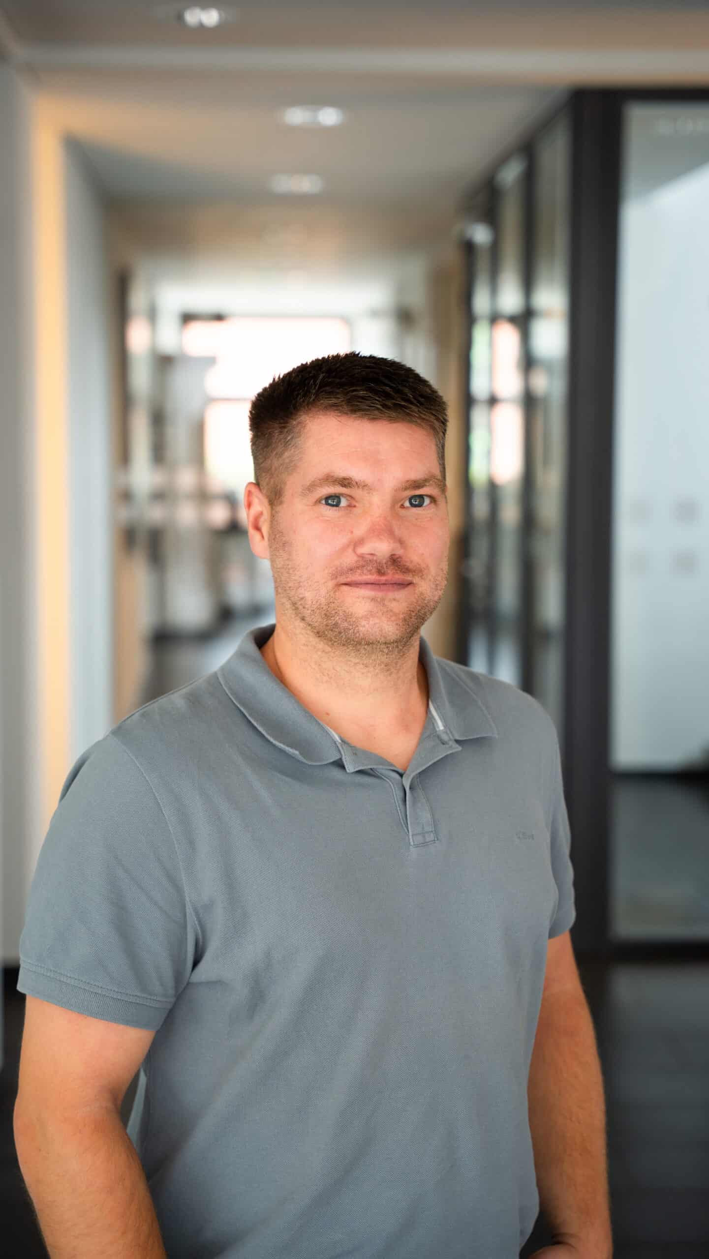 Mann mit kurzem braunem Haar und grauem Poloshirt steht in einem modernen Büroflur mit Glaswänden, neutraler Hintergrund mit weichem Licht, professionelle Atmosphäre passend zu IT-Beratung und Managed Services bei dierckgroup.