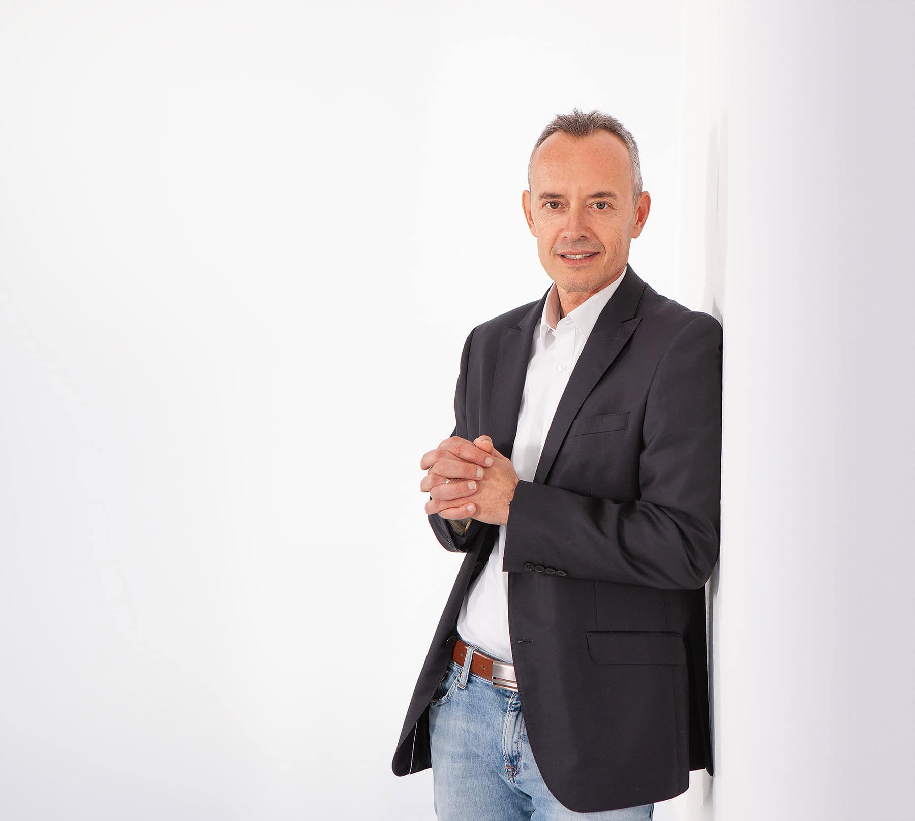 Mann mittleren Alters mit kurzem Haar, weißem Hemd, dunklem Sakko und Jeans lehnt an weißer Wand und schaut freundlich in die Kamera.