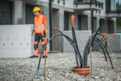Mehrere dicke Hochspannungskabel ragen aus einem Rohr im Boden einer Baustelle, im Hintergrund steht ein Elektriker mit Schutzhelm und Warnweste vor einem modernen Gebäude.