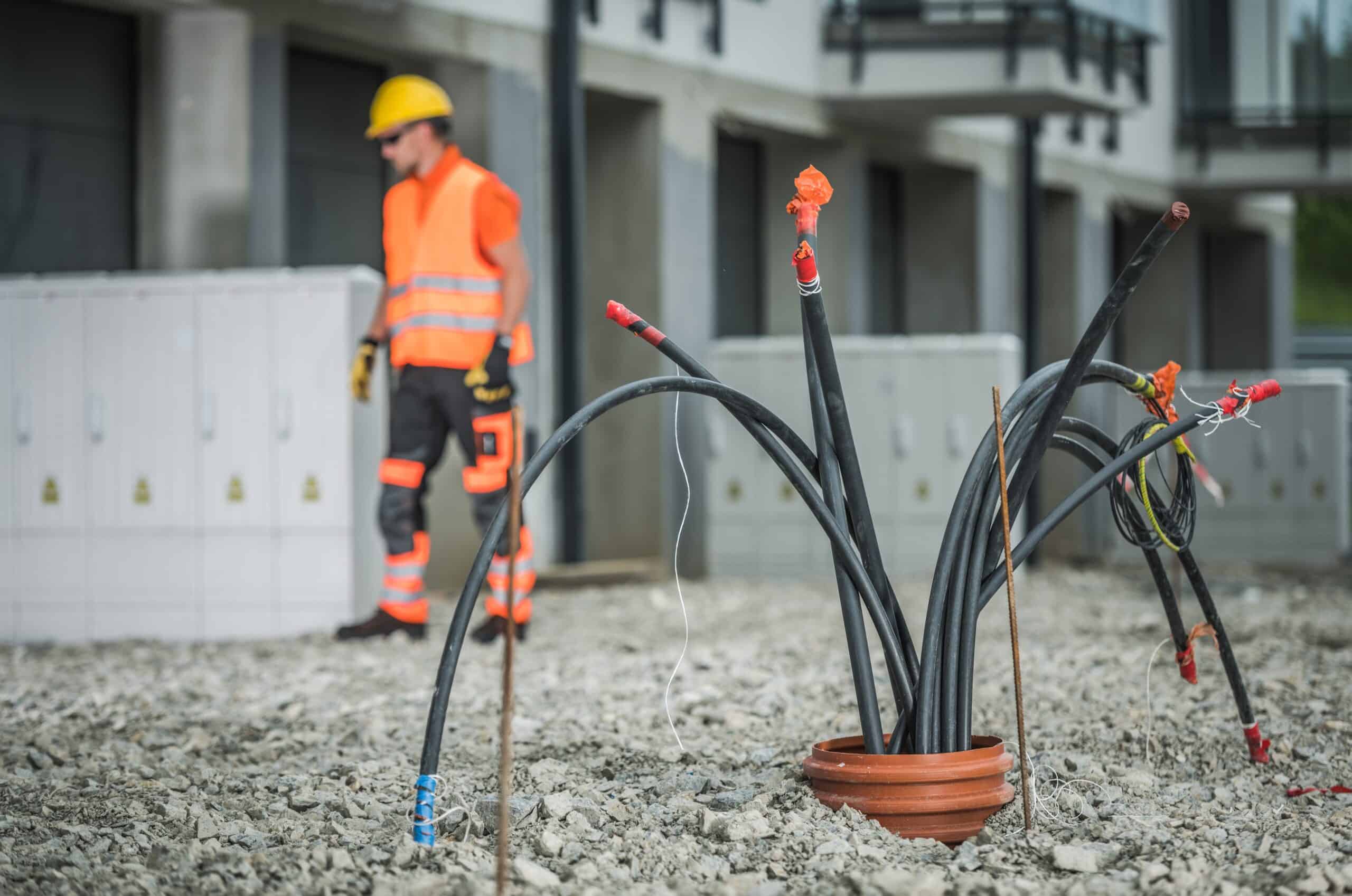 Mehrere dicke Hochspannungskabel ragen aus einem Rohr im Boden einer Baustelle, im Hintergrund steht ein Elektriker mit Schutzhelm und Warnweste vor einem modernen Gebäude.