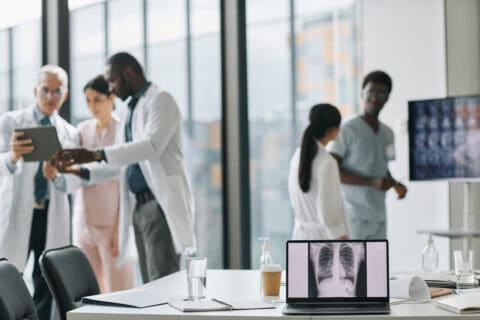 Ärzte und medizinisches Fachpersonal in einem modernen Konferenzraum, im Vordergrund ein Laptop mit Röntgenbild der Lunge auf dem Bildschirm, auf dem Tisch Wasser, Kaffeebecher und Desinfektionsmittel, helle, großflächige Fenster im Hintergrund.