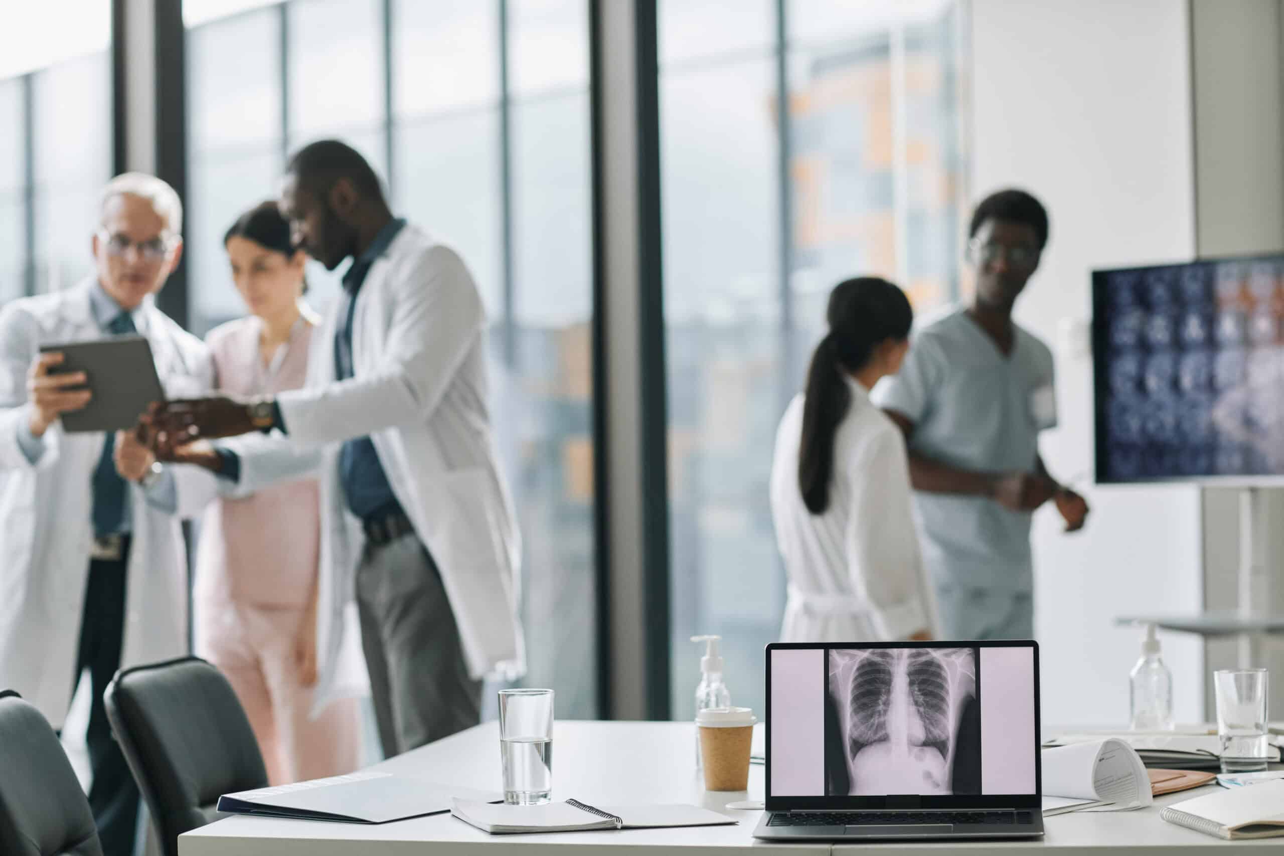 Ärzte und medizinisches Fachpersonal in einem modernen Konferenzraum, im Vordergrund ein Laptop mit Röntgenbild der Lunge auf dem Bildschirm, auf dem Tisch Wasser, Kaffeebecher und Desinfektionsmittel, helle, großflächige Fenster im Hintergrund.