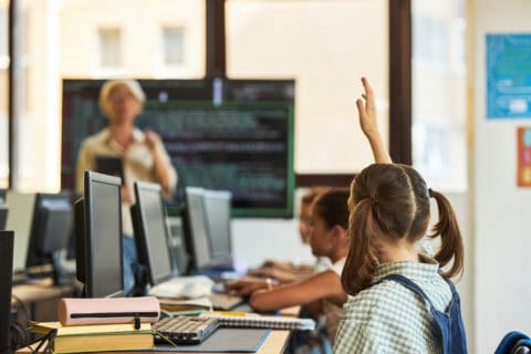 Schülerin mit Zöpfen sitzt an einem Computerarbeitsplatz in einem Klassenraum und hebt die Hand, im Hintergrund Lehrerin vor digitalem Bildschirm, weitere Kinder arbeiten an Computern, Lernumgebung für IT-Beratung und Digitalisierung.
