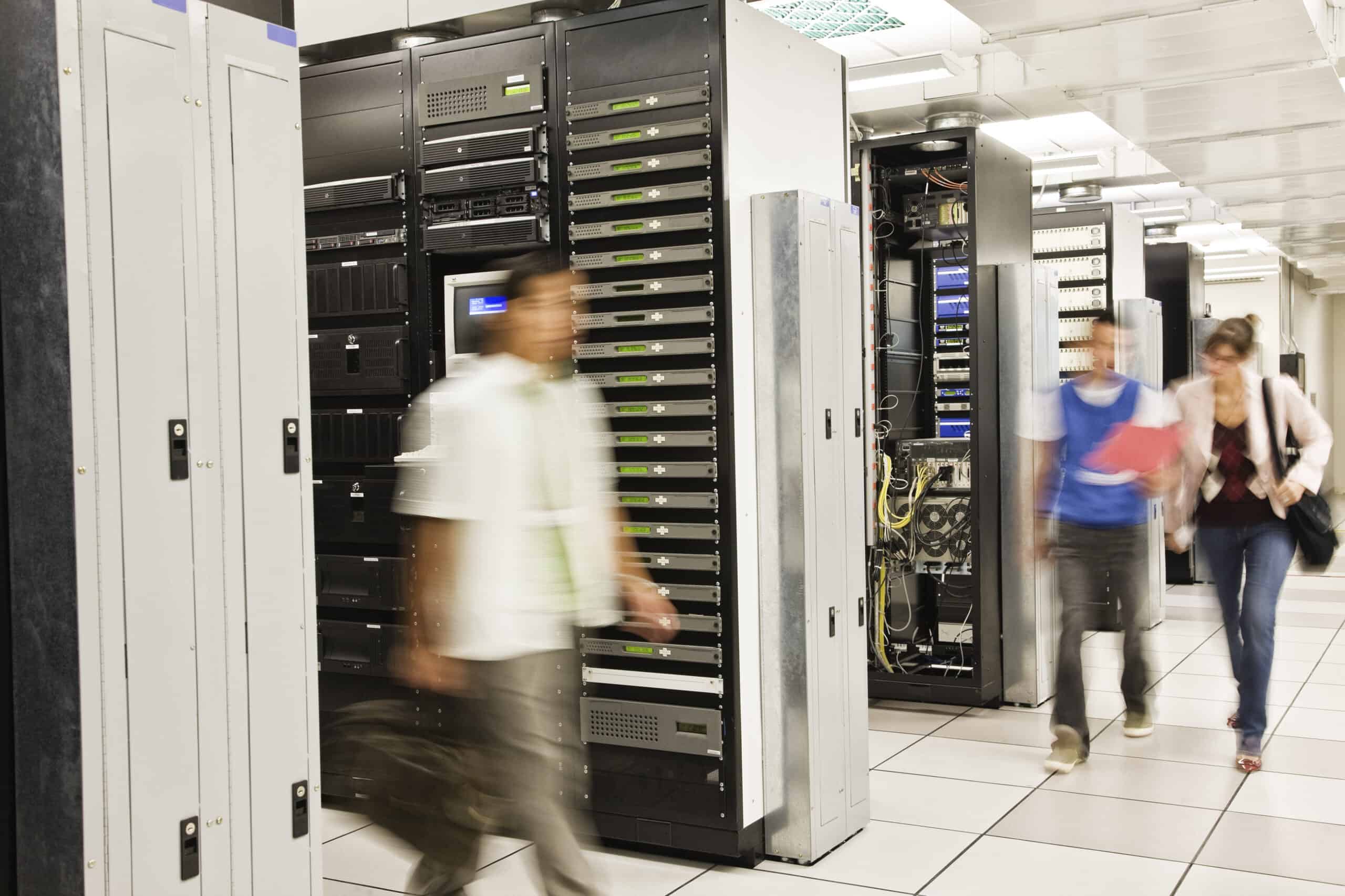 Drei Techniker in einem großen Serverraum mit mehreren Server-Racks, die an IT-Infrastruktur arbeiten, symbolisieren IT Solutions, Managed Services und Datacenter Solutions.
