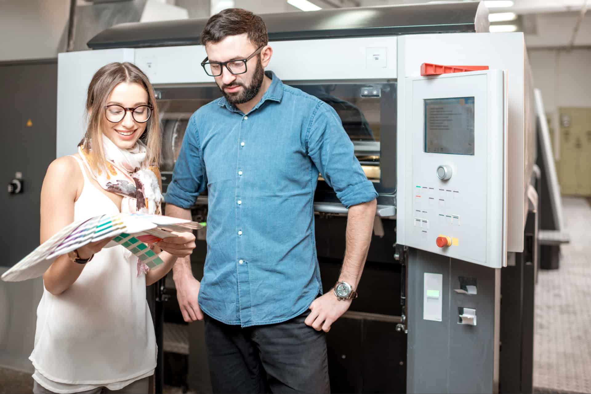 Zwei Personen mit Brille stehen vor einer großen Druckmaschine, die Frau hält einen Farbfächer und zeigt dem Mann verschiedene Farbproben, beide wirken konzentriert und im Gespräch, helle Arbeitsumgebung.