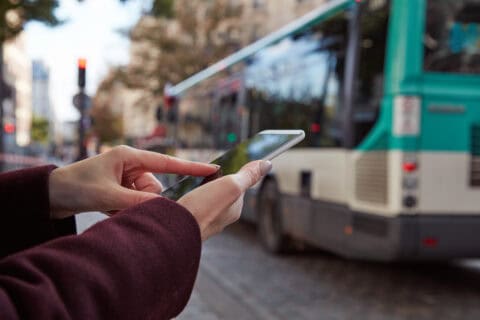 Frau hält Smartphone in der Hand und tippt darauf, im Hintergrund unscharfer Stadtbus und Straßenumgebung, symbolisiert mobile Kommunikation und digitale Vernetzung.