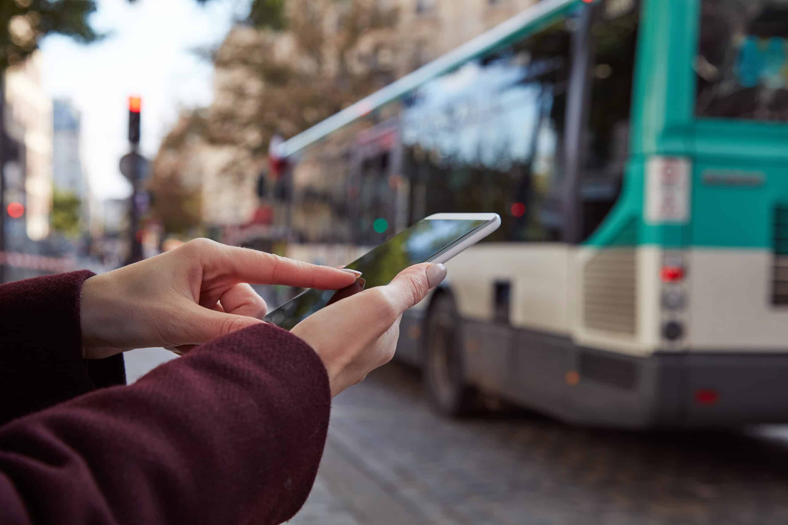 Frau hält Smartphone in der Hand und tippt darauf, im Hintergrund unscharfer Stadtbus und Straßenumgebung, symbolisiert mobile Kommunikation und digitale Vernetzung.