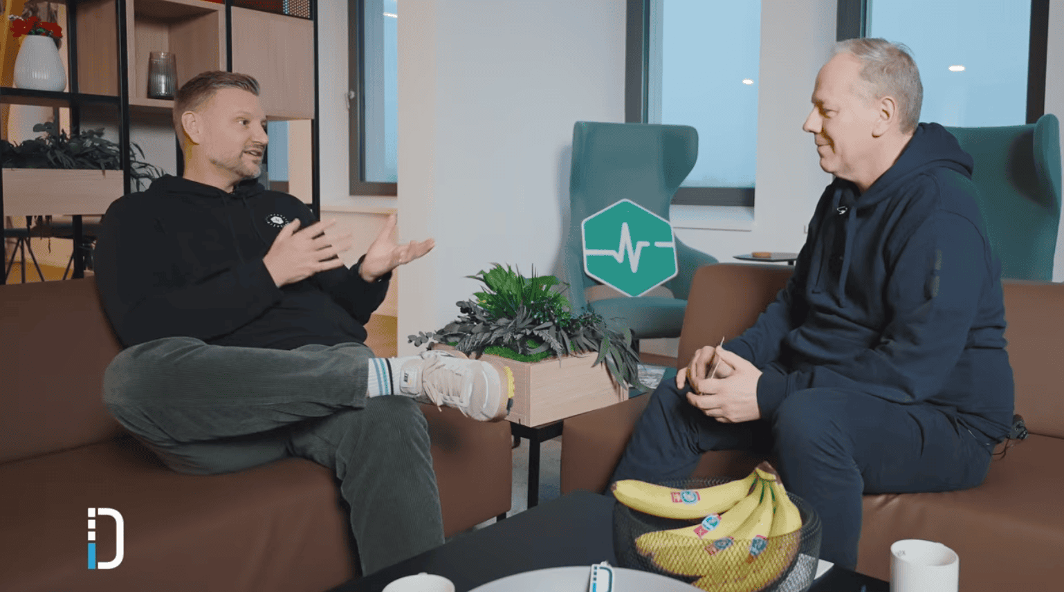 Zwei Männer in lässiger Kleidung sitzen in einem modernen Büro auf braunen Sofas und unterhalten sich. Zwischen ihnen steht ein Tisch mit einer Schale Bananen und zwei weißen Tassen. Im Hintergrund ein Regal mit Dekoration und eine grüne Sitzfläche mit einem weißen Herzschlag-Symbol. Links unten das Logo der dierckgroup.