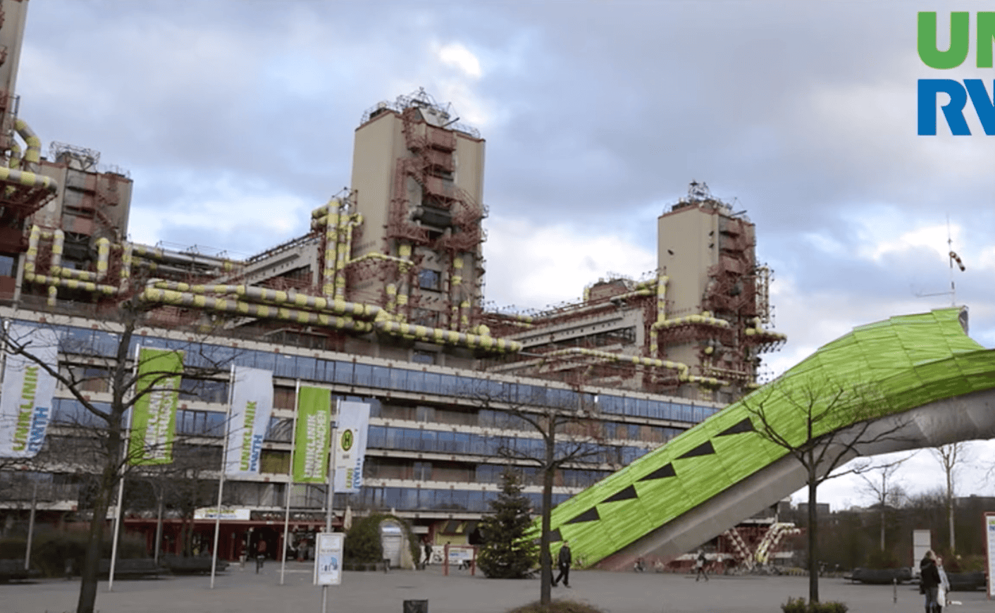 Industrielles Gebäude mit sichtbaren gelben Rohrleitungen und mehreren hohen Schornsteinen, davor ein großer grüner schräger Aufgang mit Fenstern, mehrere Fahnen mit Schrift, vereinzelte Bäume ohne Blätter, bewölkter Himmel.