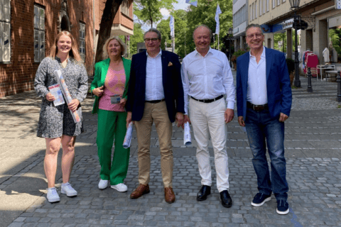 Fünf Personen stehen auf einem gepflasterten Gehweg in einer Innenstadtstraße, drei Männer und zwei Frauen, alle lächelnd und in sommerlicher Kleidung. Zwei Personen halten gerollte Pläne oder Dokumente. Im Hintergrund sind Backsteingebäude, Bäume und Ladengeschäfte zu sehen. Die Szene vermittelt ein freundliches, professionelles Teamumfeld, passend zu den Themen IT Solutions, Managed Services, IT-Beratung und Digitalisierung der dierckgroup.