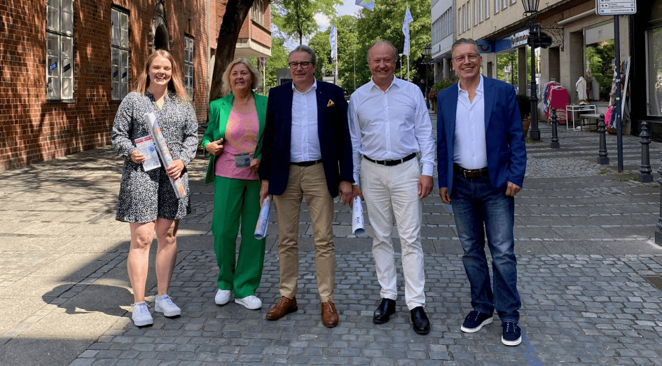 Fünf Personen stehen auf einem gepflasterten Gehweg in einer Innenstadtstraße, drei Männer und zwei Frauen, alle lächelnd und in sommerlicher Kleidung. Zwei Personen halten gerollte Pläne oder Dokumente. Im Hintergrund sind Backsteingebäude, Bäume und Ladengeschäfte zu sehen. Die Szene vermittelt ein freundliches, professionelles Teamumfeld, passend zu den Themen IT Solutions, Managed Services, IT-Beratung und Digitalisierung der dierckgroup.