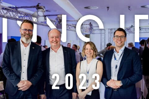 Vier Personen in Business-Kleidung stehen vor einem beleuchteten Schriftzug „INNO IT 2023“ in einem modernen Veranstaltungsraum mit weiteren Gästen im Hintergrund.