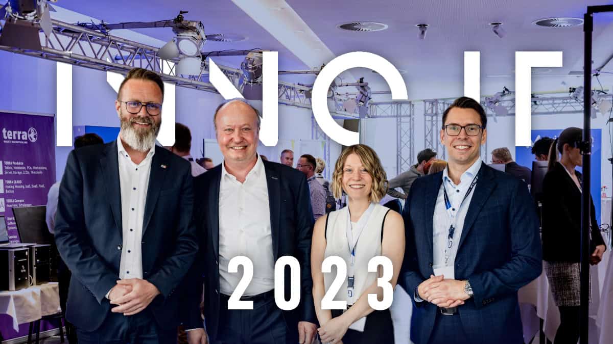 Vier Personen in Business-Kleidung stehen vor einem beleuchteten Schriftzug „INNO IT 2023“ in einem modernen Veranstaltungsraum mit weiteren Gästen im Hintergrund.