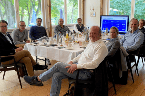 Neun Personen sitzen an einem rechteckigen Tisch in einem hellen Raum mit großen Fenstern und Blick ins Grüne. Auf dem Tisch stehen Wasserflaschen, Gläser, Tassen und Unterlagen. Im Hintergrund ist ein Bildschirm mit einer Präsentation zur LinkedIn-Kampagne der dierckgroup zu sehen. Die Szene vermittelt eine Besprechung oder ein Boardmeeting, passend zu IT-Beratung, Managed Services und Digitalisierung im Kontext der dierckgroup.
