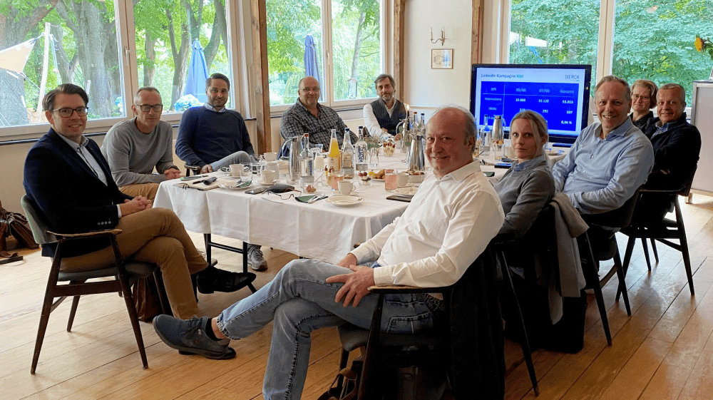 Neun Personen sitzen an einem rechteckigen Tisch in einem hellen Raum mit großen Fenstern und Blick ins Grüne. Auf dem Tisch stehen Wasserflaschen, Gläser, Tassen und Unterlagen. Im Hintergrund ist ein Bildschirm mit einer Präsentation zur LinkedIn-Kampagne der dierckgroup zu sehen. Die Szene vermittelt eine Besprechung oder ein Boardmeeting, passend zu IT-Beratung, Managed Services und Digitalisierung im Kontext der dierckgroup.