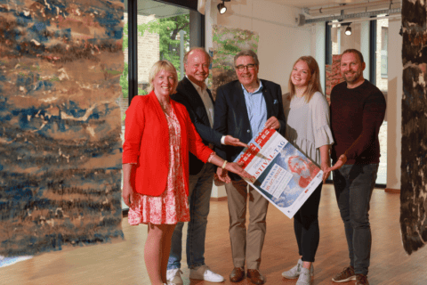 Fünf Personen in einem hellen Raum mit großen Fenstern und abstrakten, farbigen Textilkunstwerken an der Decke. Drei Männer und zwei Frauen stehen nebeneinander, lächeln und halten gemeinsam ein Plakat mit dem Schriftzug „Festival“. Die Atmosphäre wirkt freundlich und kooperativ.