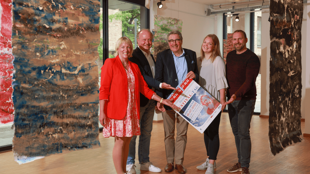 Fünf Personen in einem hellen Raum mit großen Fenstern und abstrakten, farbigen Textilkunstwerken an der Decke. Drei Männer und zwei Frauen stehen nebeneinander, lächeln und halten gemeinsam ein Plakat mit dem Schriftzug „Festival“. Die Atmosphäre wirkt freundlich und kooperativ.
